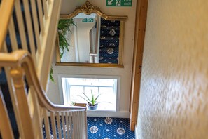 Un escalier avec une rampe en bois, une plante en pot sur le palier, un miroir au mur et un panneau indiquant la « sortie de secours ».