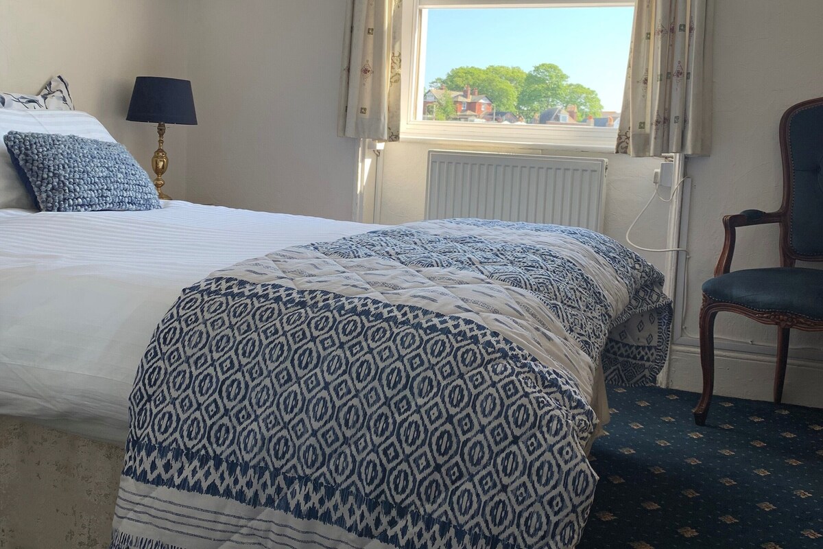 Une chambre d’hôtel avec un lit, une chaise, une fenêtre avec des rideaux, un radiateur et une étagère ornée d’objets décoratifs.