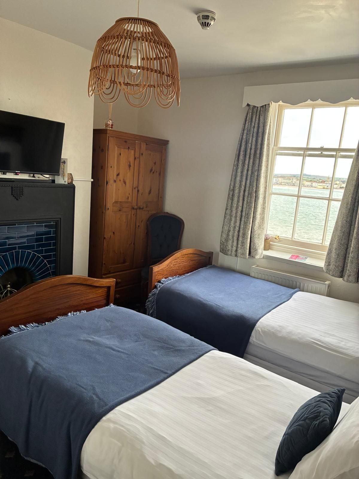 A room with two beds, a wooden wardrobe, a television on a stand, and a window with a view of the sea.
