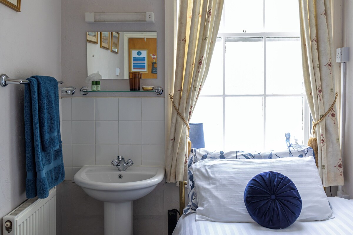 Une chambre à coucher avec un lit, une fenêtre avec des rideaux, une table de chevet et une salle de bain avec un lavabo et un porte-serviettes.