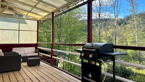 Une terrasse couverte équipée d’un barbecue, de meubles en osier et offrant une vue sur une zone boisée.