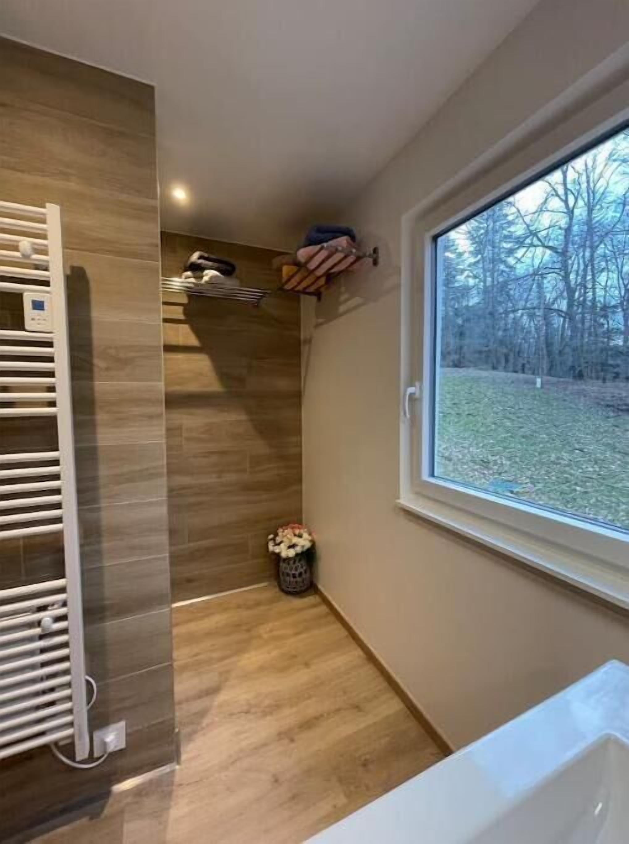 A bathroom with a bathtub, a window with a view of trees, a towel rack, and a trash bin.
