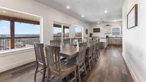 A spacious dining area with a long table and multiple chairs, large windows with a view, and a kitchen area in the background.
