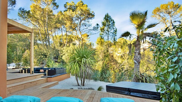 Un espace extérieur moderne, aménagé avec une terrasse en bois et des coussins bleus, entouré d’une végétation luxuriante et d’arbres.