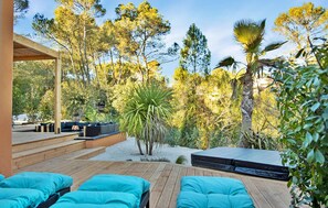 A modern outdoor seating area with wooden deck and blue cushions, surrounded by lush greenery and trees. - Les Villas Au Palmier (Cannes-Et-Clairan)