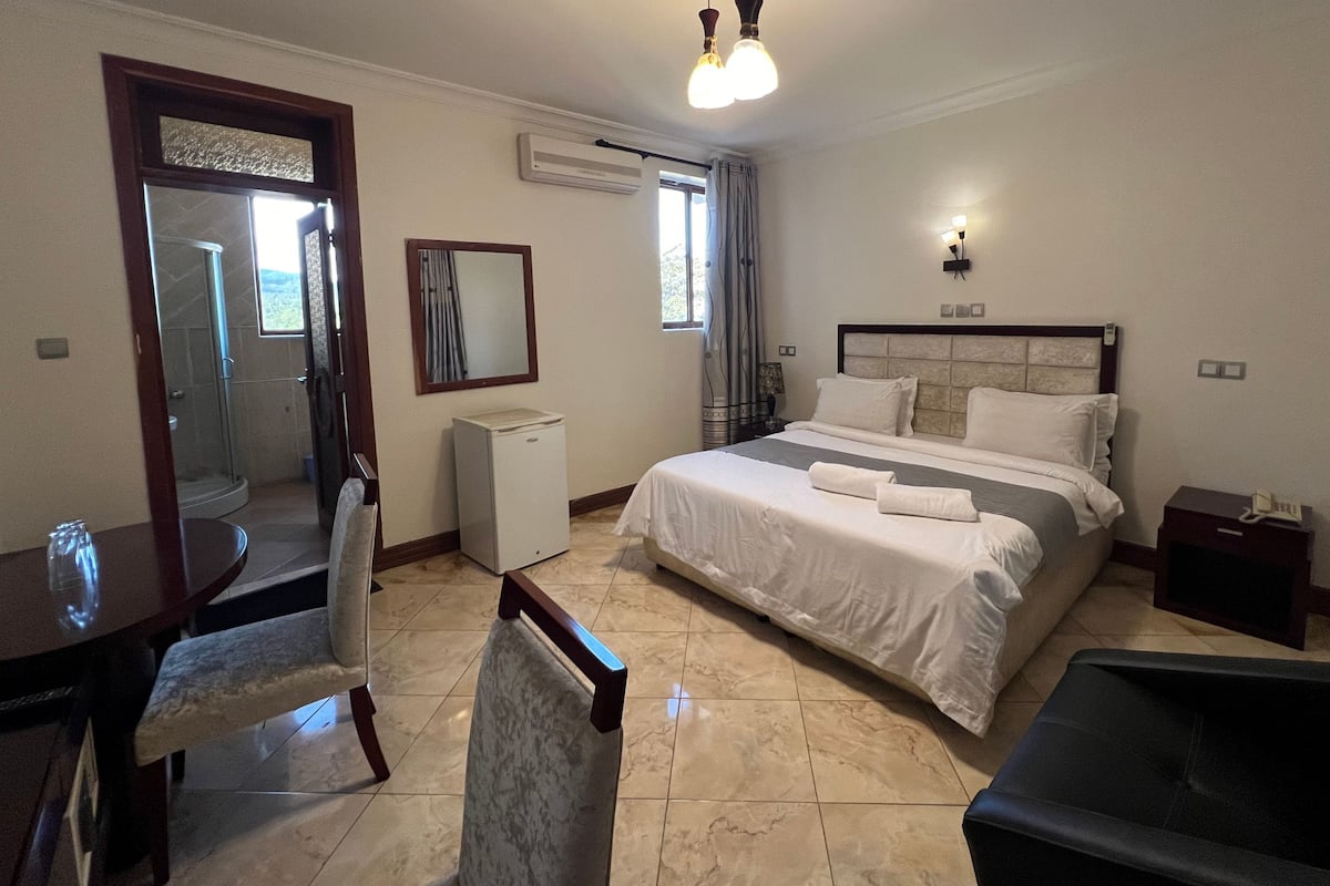 A hotel room with a bed, a desk, a chair, a small refrigerator, a mirror, and a window with curtains.