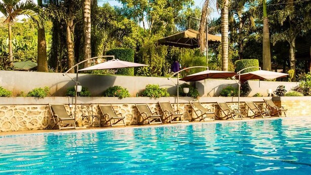 A swimming pool with lounge chairs and umbrellas, surrounded by palm trees and greenery. - Nejobugg palace hotel (Arusha)