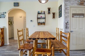 Une table à manger rustique en bois, accompagnée de six chaises, une étagère en bois sur laquelle sont disposés divers objets, et une porte avec une pancarte.