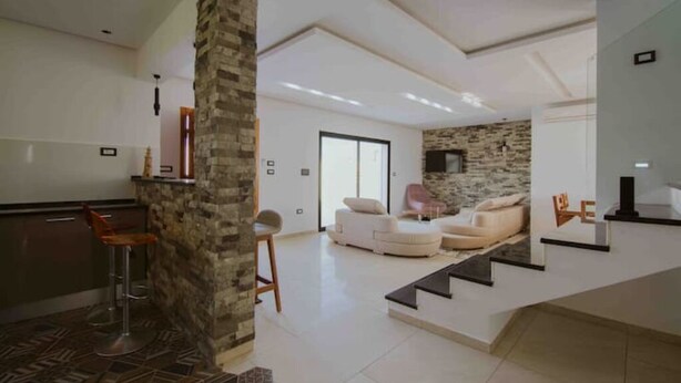 A modern living room with a stone pillar, a white sofa, and a staircase leading to another level. - Djerba Villa Piscine Prestige (Temlel)