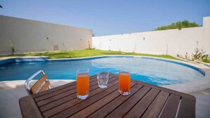 A poolside table with three glasses of orange beverage, a pool, and a white wall in the background. - Djerba Villa Piscine Prestige (Temlel)
