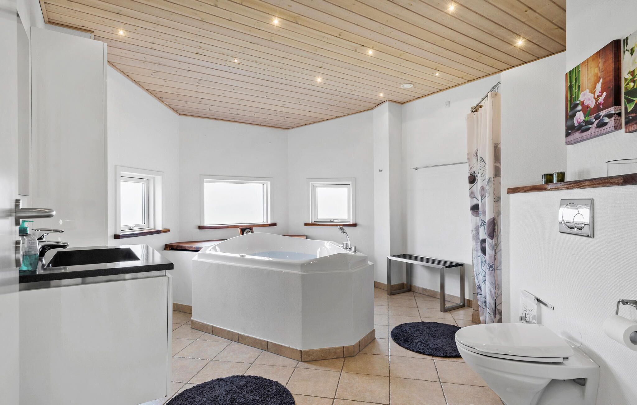 Un baño moderno con una bañera independiente, un inodoro y un lavabo. Hay tres ventanas que aportan luz natural, y una zona de ducha con cortina.