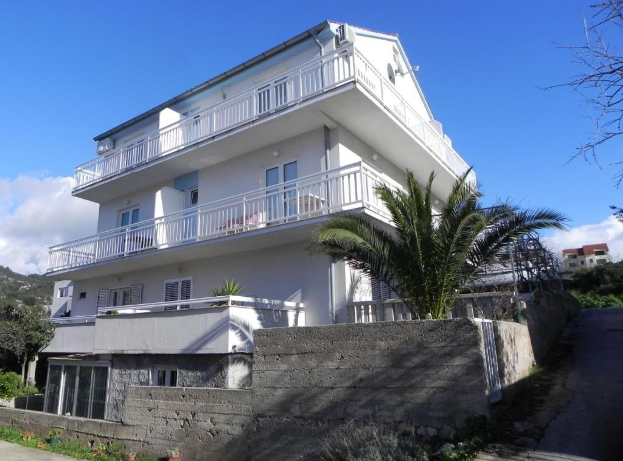 A multi-story building with balconies, a stone retaining wall, and a palm tree.