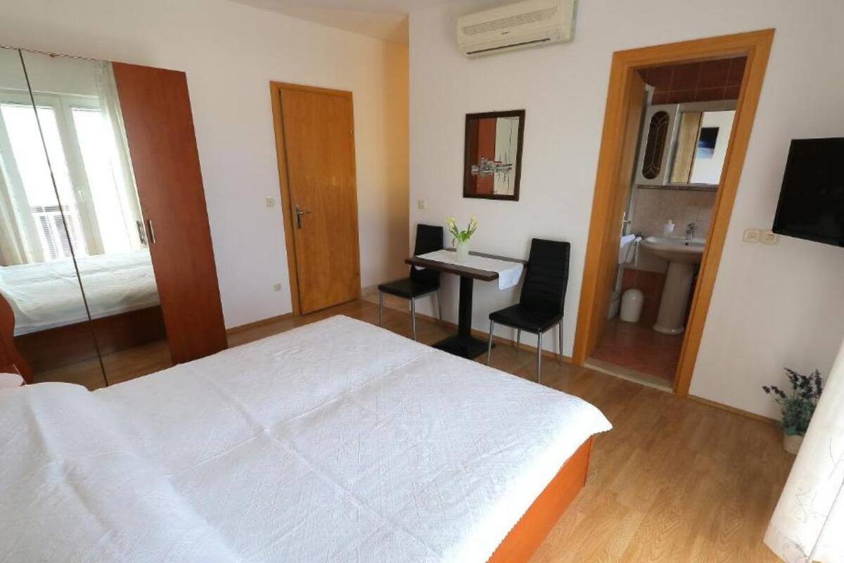 A hotel room with a large bed, a desk with a chair, a small table with a vase, a mirror, and a TV.
