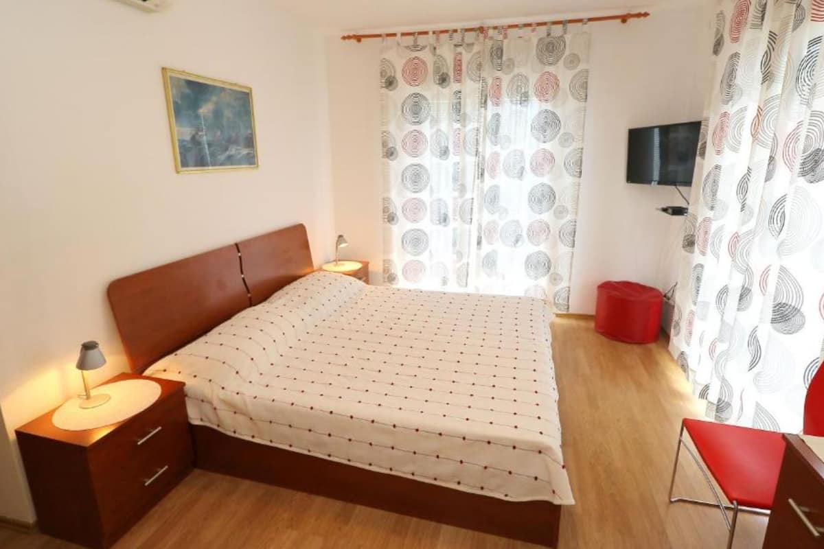 A bedroom with a bed, a nightstand, a lamp, a TV, and a red chair.