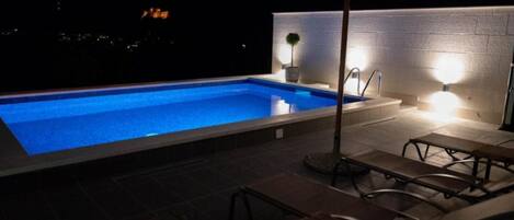 Piscina exterior com um deck ladrilhado, um banco e um guarda-sol.