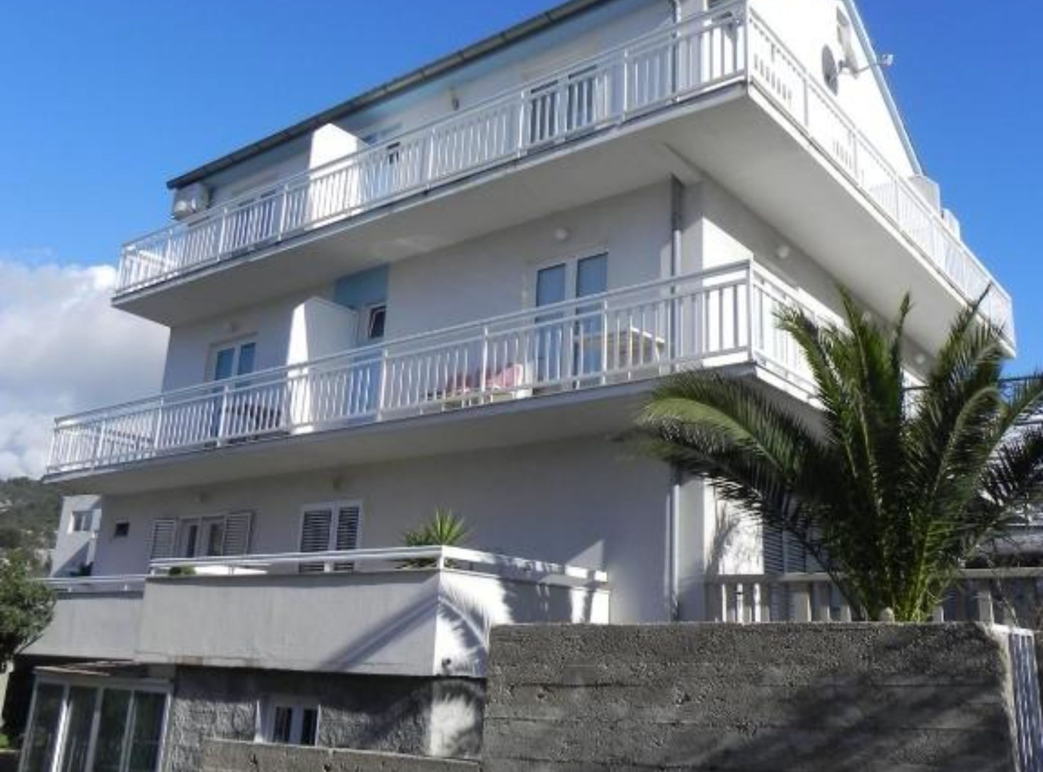 Un edificio a più piani con balconi, un muro in pietra in primo piano e un cielo azzurro limpido.