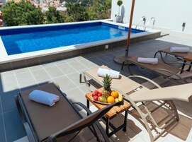 A rooftop pool area with lounge chairs, a table with fruit, and a view of the city.