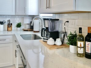 Une cuisine moderne avec des meubles blancs, un évier en acier inoxydable, une machine à café, un mixeur et deux bouteilles de vin.