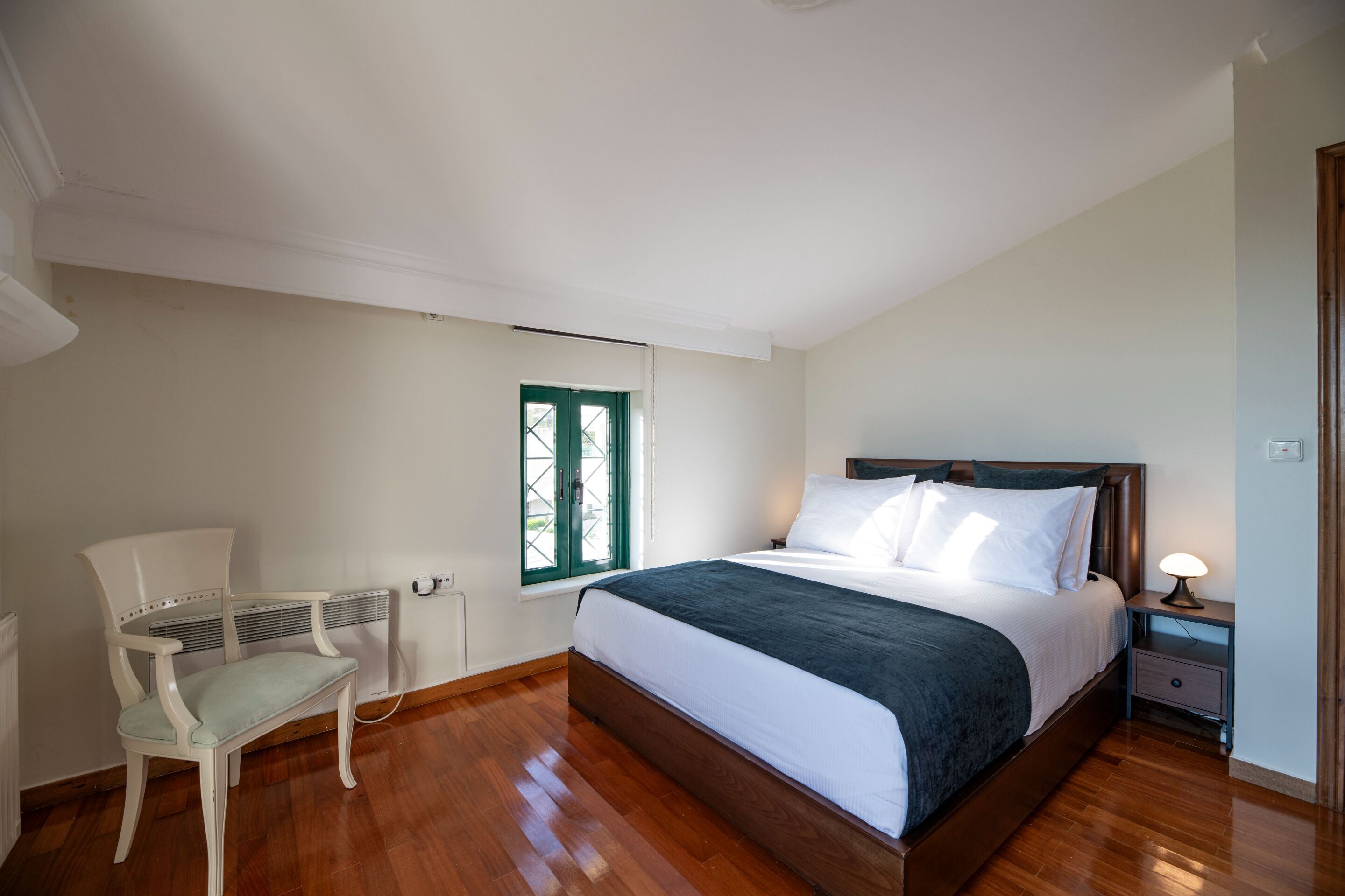 A bedroom with a wooden bed, a chair, a nightstand, and a window with green shutters.