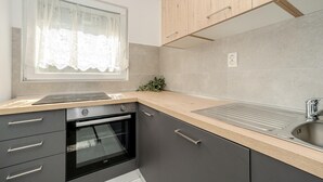 Une cuisine moderne avec des meubles gris, un four encastré, un évier en acier inoxydable et une fenêtre ornée de rideaux en dentelle.