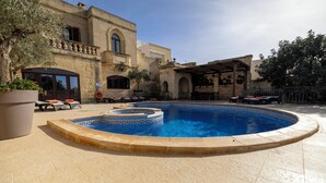 Una piscina con solárium de piedra, mobiliario exterior y un edificio con ventanas arqueadas y balcón.