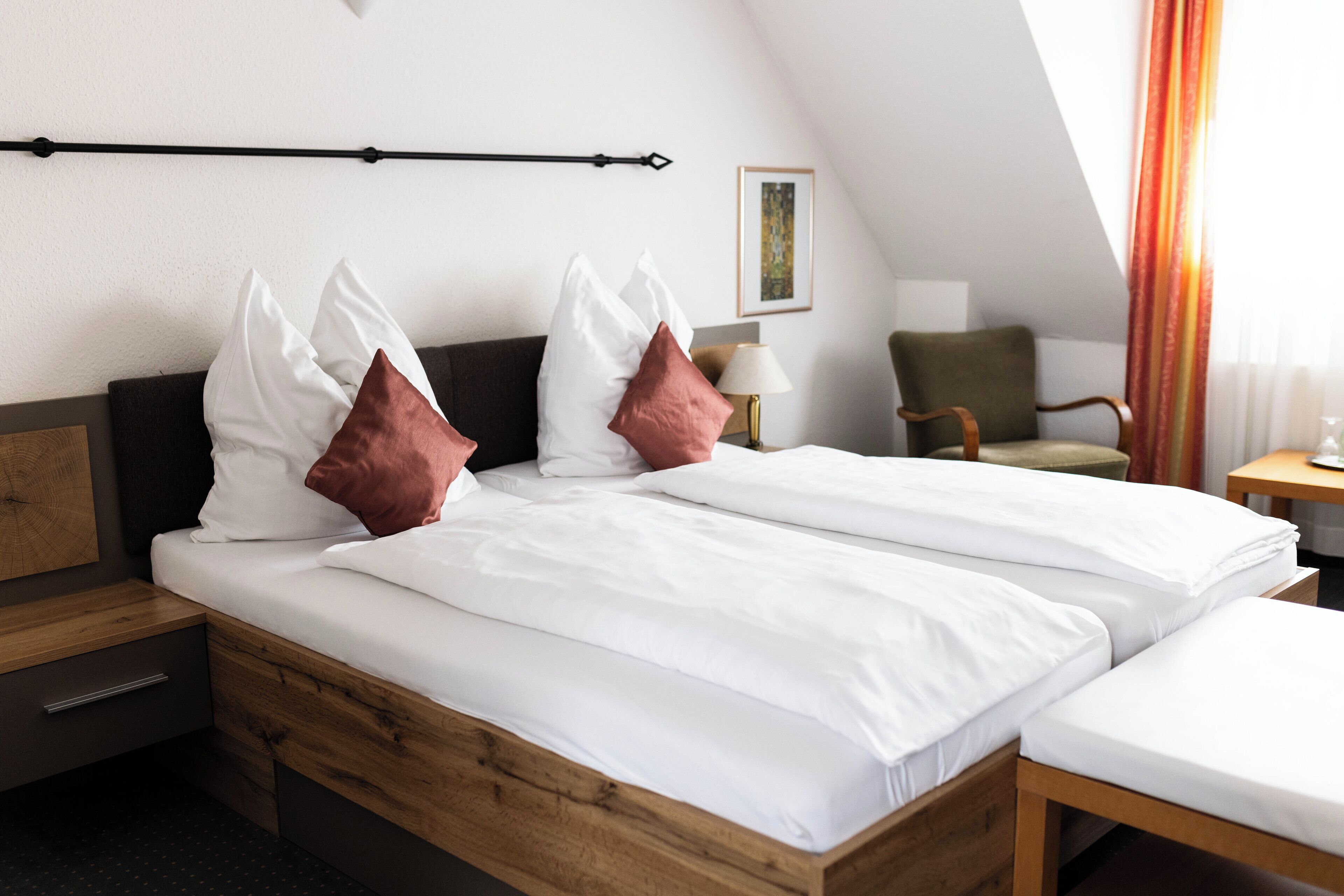 A double bed with white bedding and brown pillows, a wooden nightstand, a chair, and a small table with a lamp.