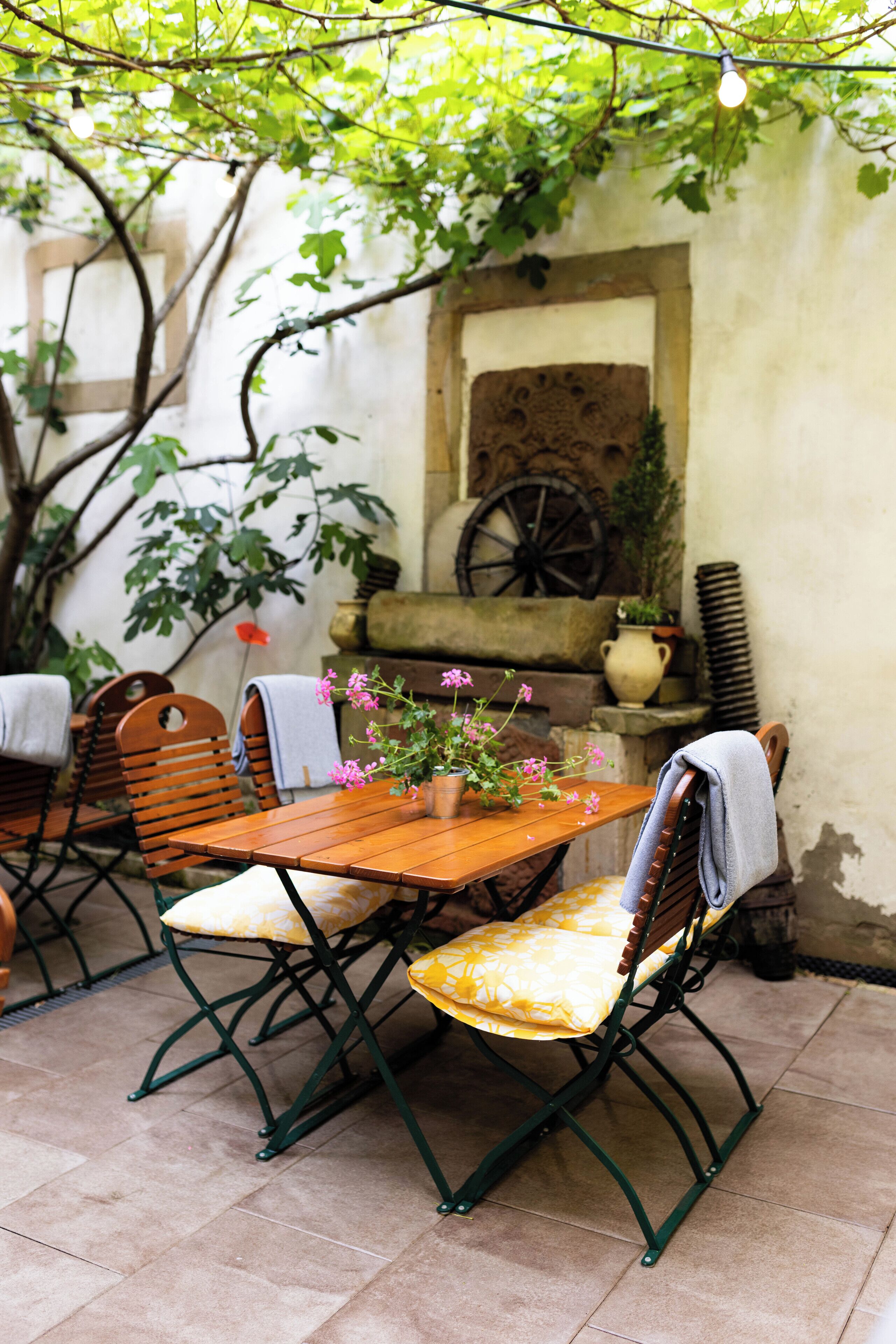 An outdoor patio with a wooden table and chairs, surrounded by greenery and a stone wall.