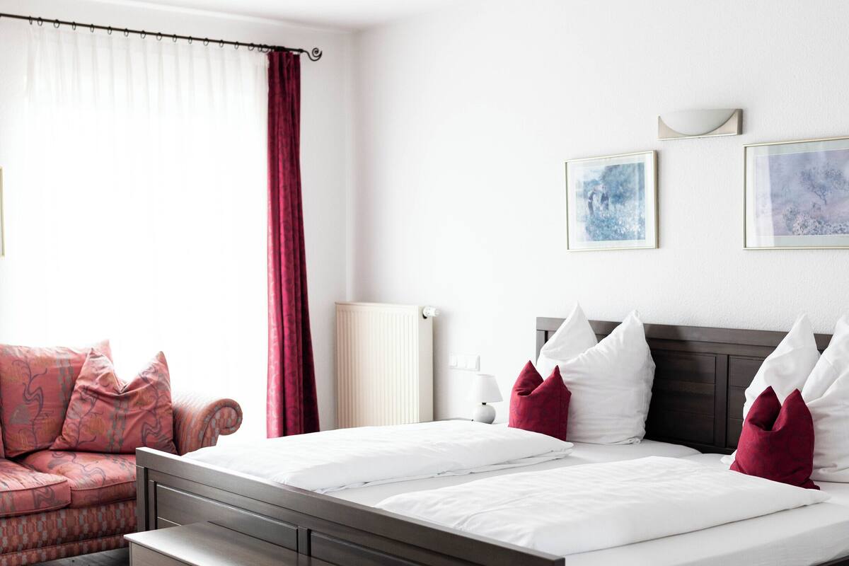Ein modernes Hotelzimmer mit einem großen Bett, einer Sitzbank am Fußende, einer Couch und einem Fenster mit Vorhängen.