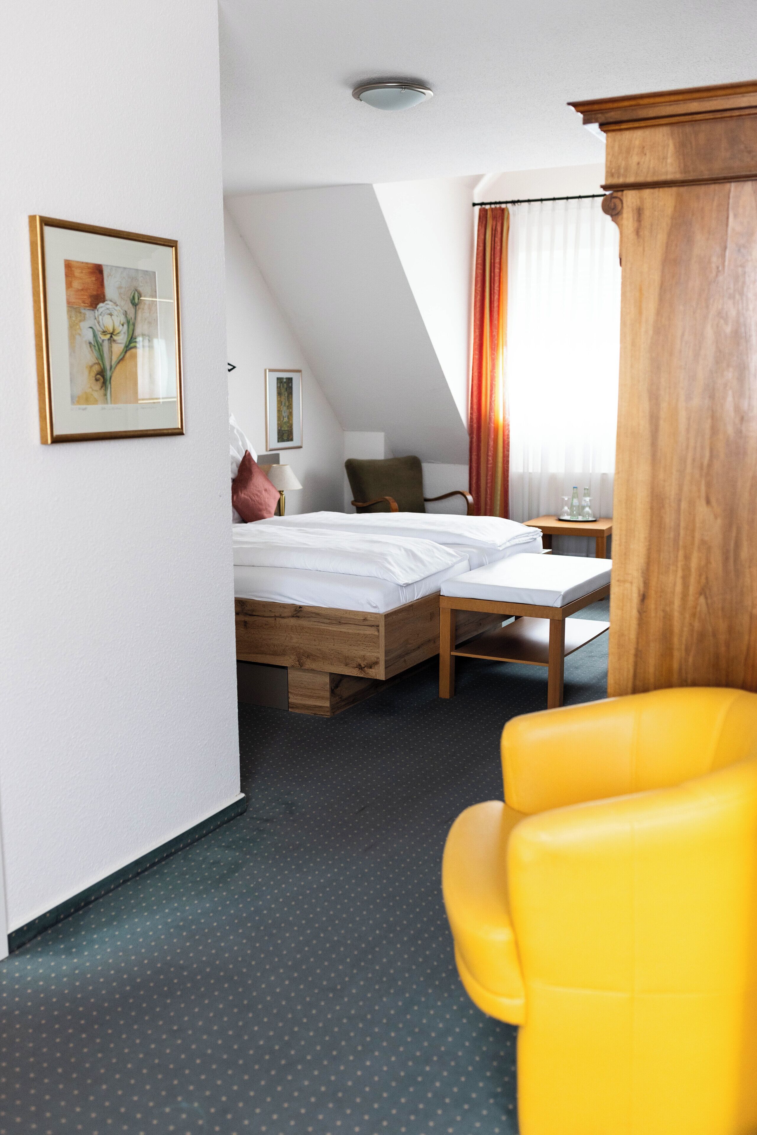 A hotel room with a bed, a wooden nightstand, a yellow chair, and a window with curtains.