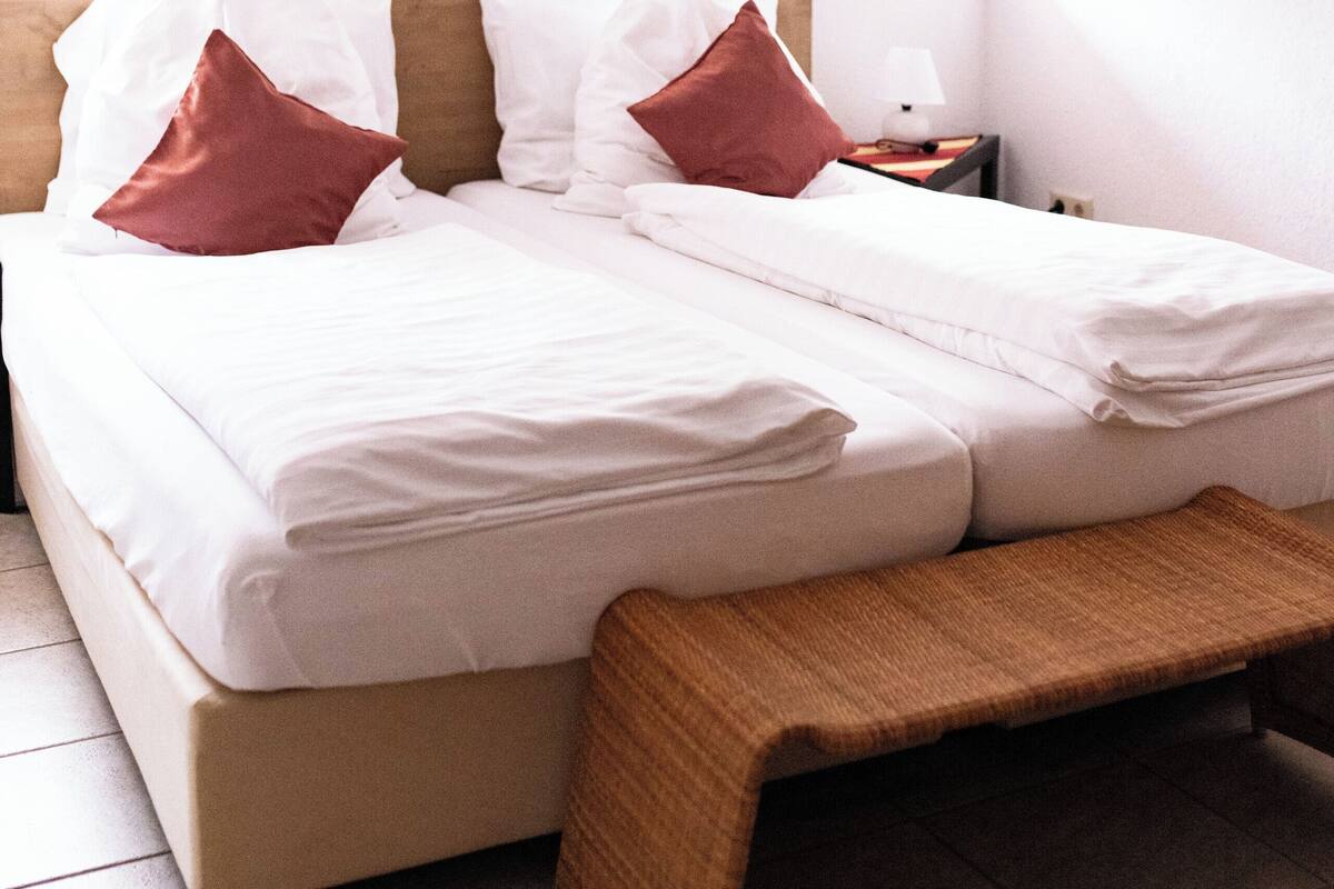 Ein ordentlich bezogenes Bett mit weißen Leinen und zwei roten Kissen, eine Holzbank am Fußende des Bettes, ein kleiner Nachttisch mit Lampe sowie ein farbenfrohes abstraktes Gemälde an der Wand.
