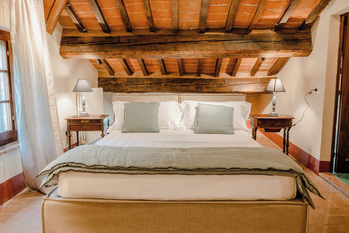 A bedroom with a wooden ceiling, a bed with white linens, two bedside tables with lamps, and a window with curtains.