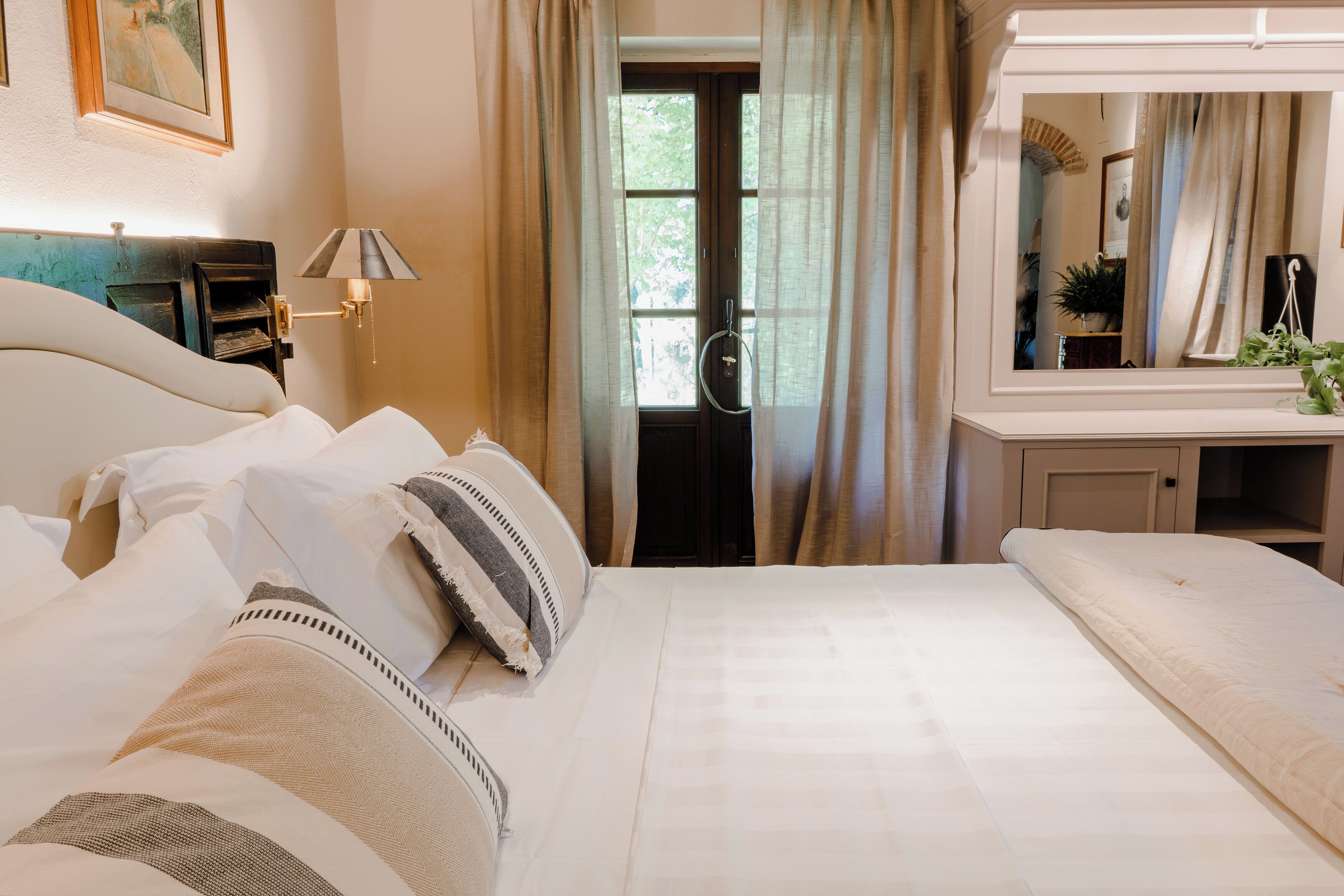 A hotel room with a large bed, a bedside table with a lamp, a mirror, and a view of the outdoors through a window.
