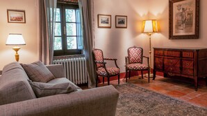 Un salon comprenant un canapé, deux fauteuils, une lampe sur pied, un radiateur, une fenêtre avec des rideaux, un meuble et des tableaux encadrés accrochés au mur.