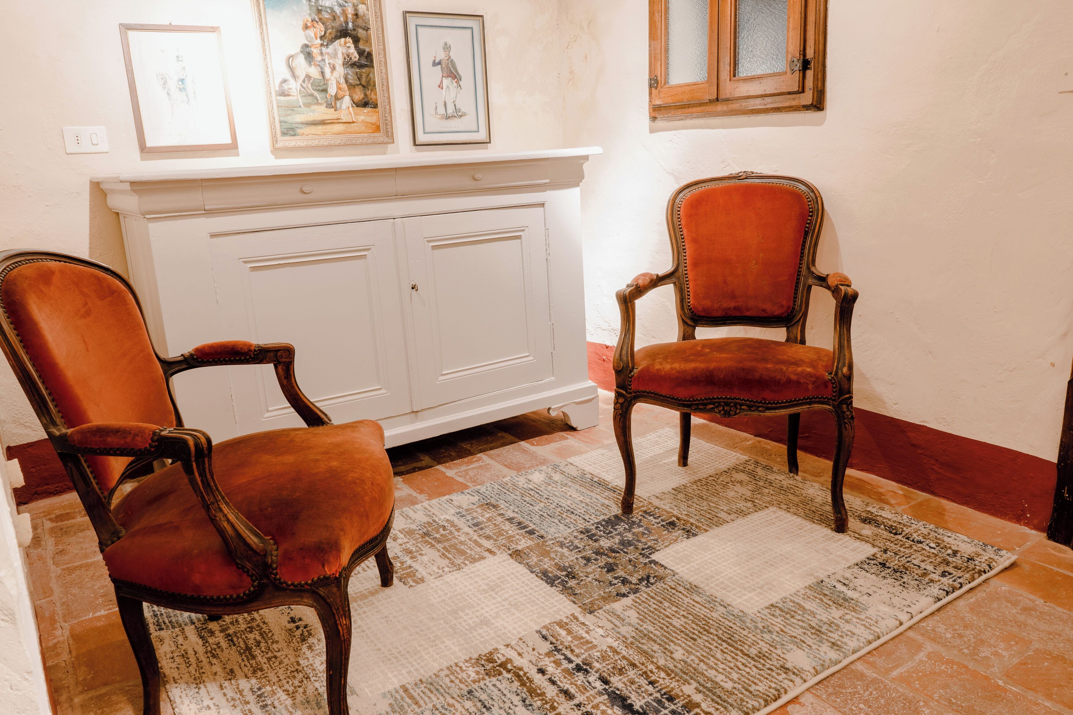 Two antique chairs with velvet upholstery, a white cabinet with artwork above, and a patterned rug on a tiled floor.