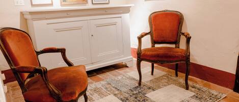 Two antique chairs with velvet upholstery, a white cabinet with artwork above, and a patterned rug on a tiled floor.