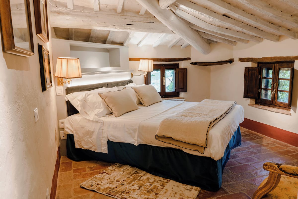 A bedroom with a bed, bedside lamps, a window, and a rustic wooden ceiling.