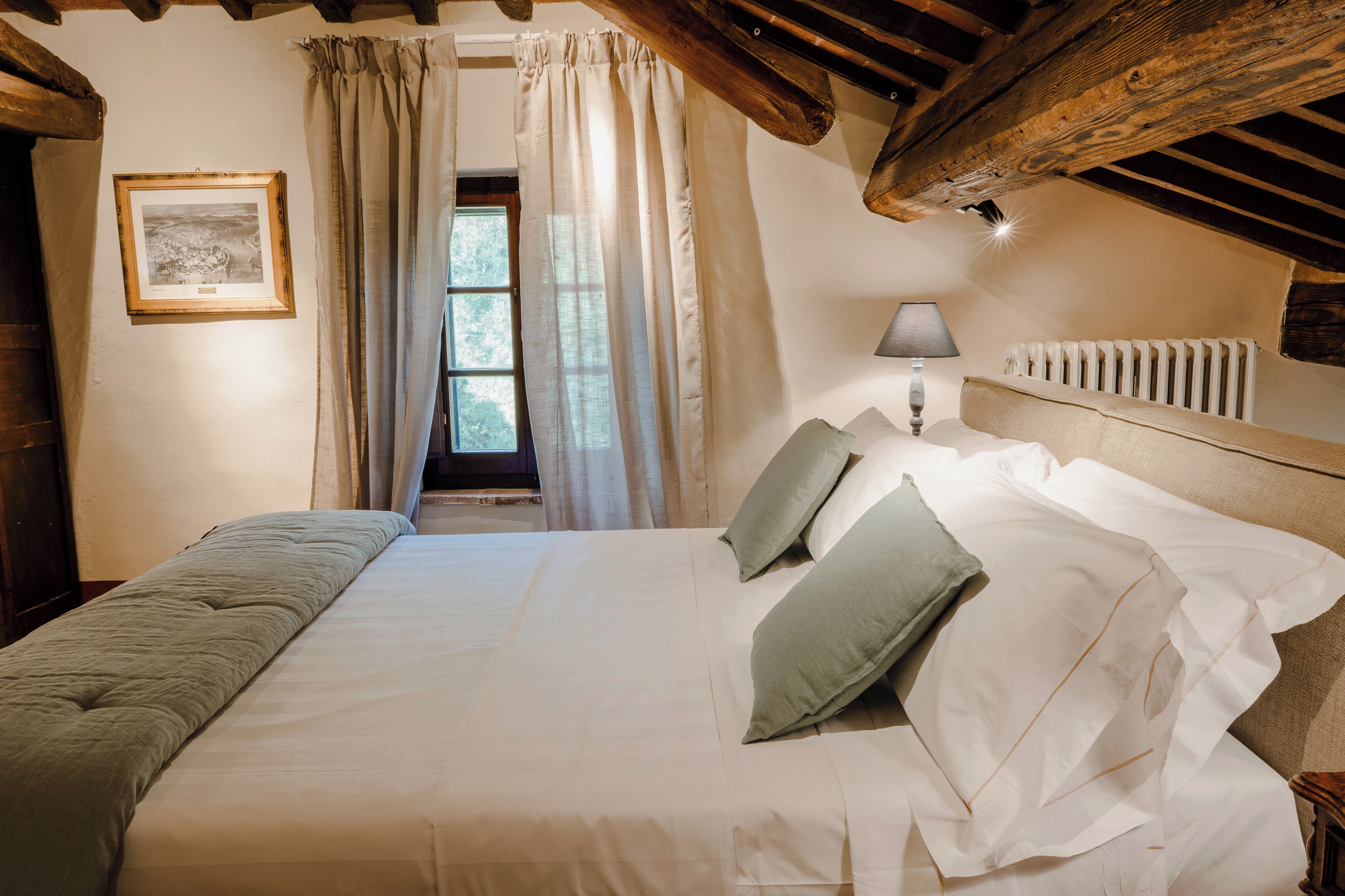 A bedroom with a large bed, a nightstand, a lamp, a framed picture on the wall, and a window with curtains.