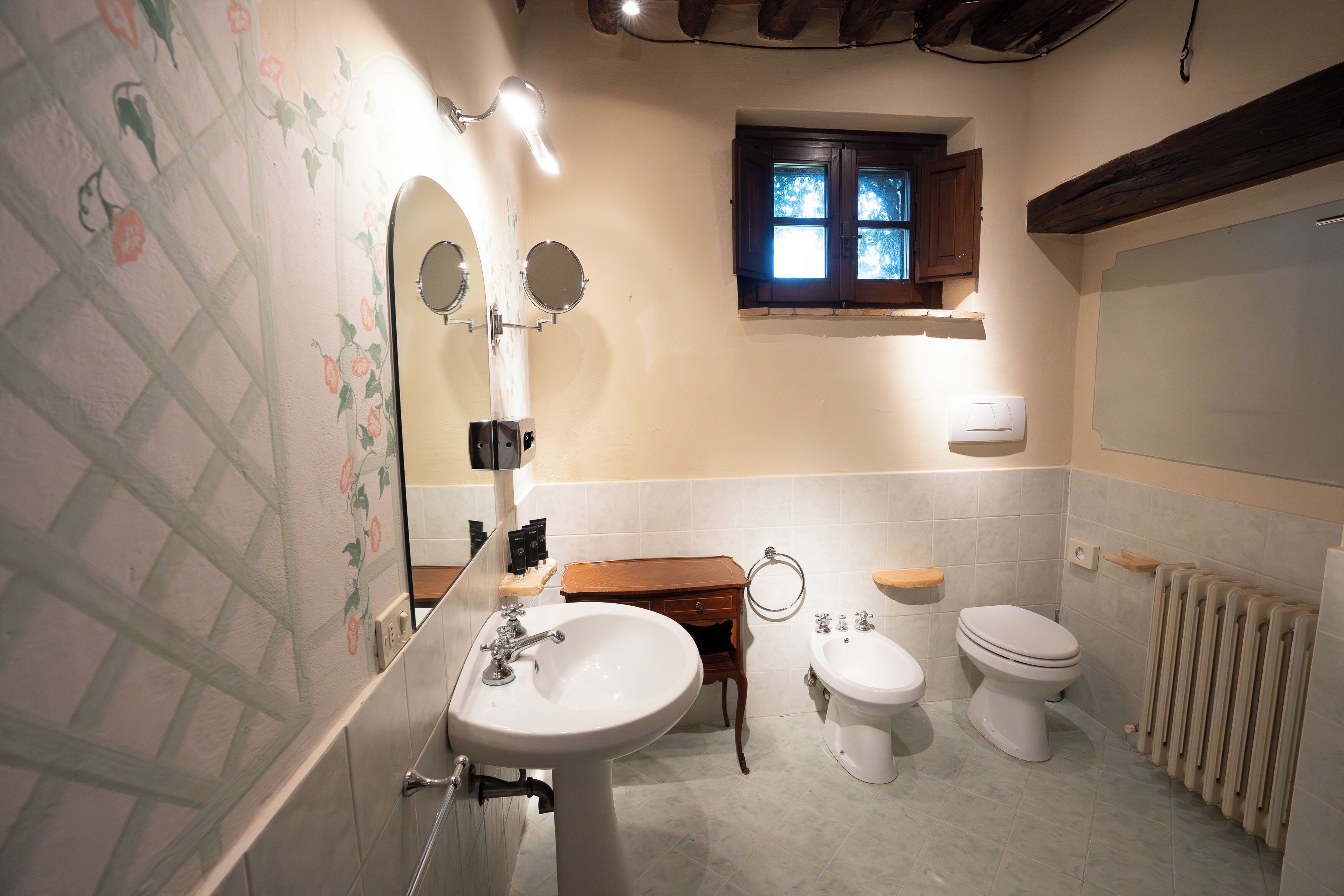 Un baño con lavamanos, dos inodoros, un armario de madera y ventana con vistas.