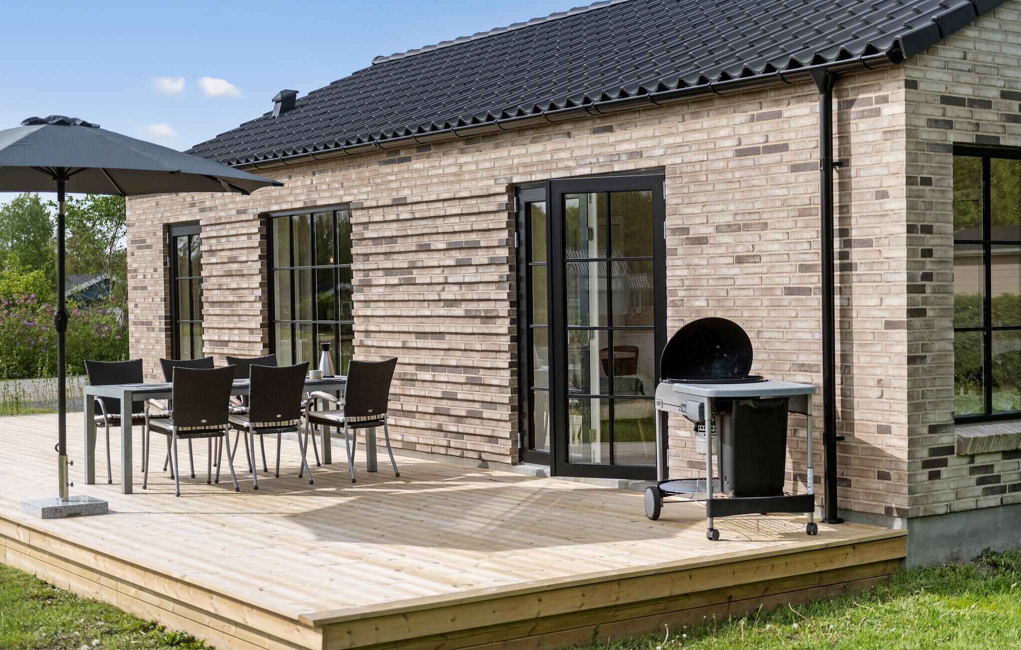 Une maison moderne avec une façade en briques, de grandes fenêtres et une terrasse en bois aménagée avec du mobilier d’extérieur.