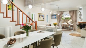 A modern living room with a white sofa, dining table, and a staircase. There are several paintings on the walls and a vase with flowers on the table. - Villa Ithaca.  Country side villa close to all worth visiting sights in Chania.  (CHANIA)