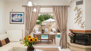 A living room with a sofa, a glass coffee table, a fireplace, and a view of an outdoor pool area. - Villa Ithaca.  Country side villa close to all worth visiting sights in Chania.  (CHANIA)