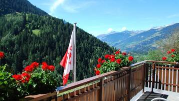 A balcony with a wooden railing overlooking a mountainous landscape, a flag, and red flowers.