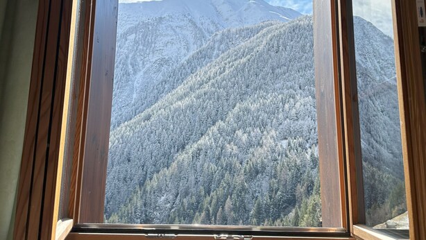 A wooden-framed window with a view of snow-covered mountains and forested hills. - Jägerheim (Ernen)