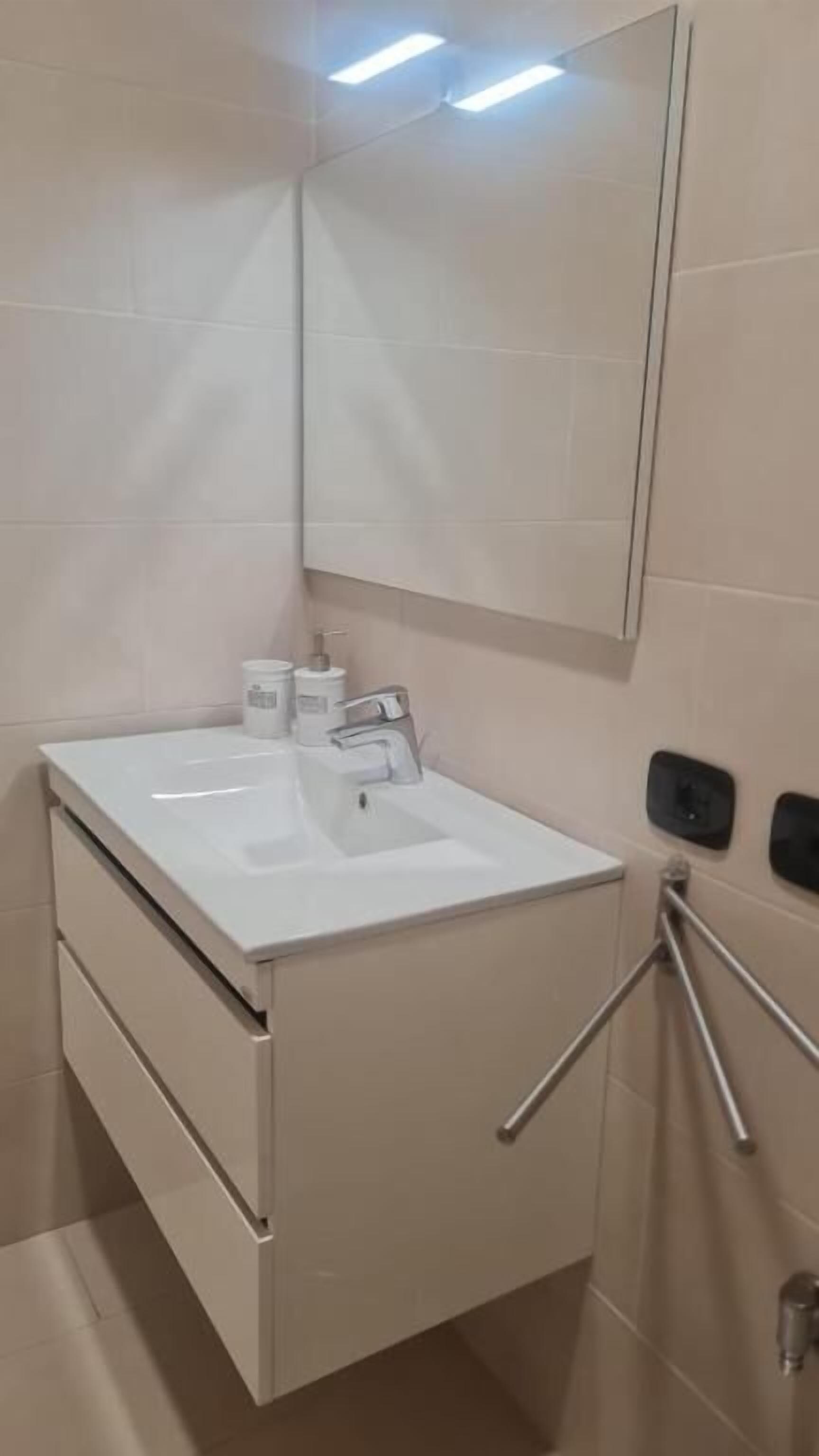 Een moderne badkamer met een wit wastafelmeubel, spiegel en opbergkast. Op het aanrecht staan twee handdrogers en een zeepdispenser.