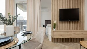 Una habitación de hotel moderna con una mesa de vidrio, un sillón cómodo, un televisor de pantalla plana montado en una pared de paneles de madera y una cama con cabecera blanca.