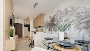 Un espacio de sala moderno con comedor que incluye una mesa y sillas de vidrio, una cocina con armarios de madera y un mural de palmeras en la pared.
