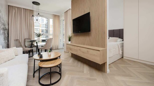 Una habitación de hotel moderna con una televisión de pantalla plana montada en una pared revestida de madera, una cama con cabecero oscuro, una zona de estar con una mesa redonda y sillas, y un ventanal con vistas a la ciudad.