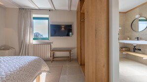 Une chambre à coucher comprenant un lit, une fenêtre, un téléviseur, un banc et une porte en bois donnant sur une salle de bain équipée d’un lavabo et d’un miroir.