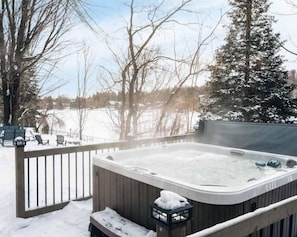 Un bain à remous sur une terrasse en bois, avec de la neige au sol et des arbres en arrière-plan.