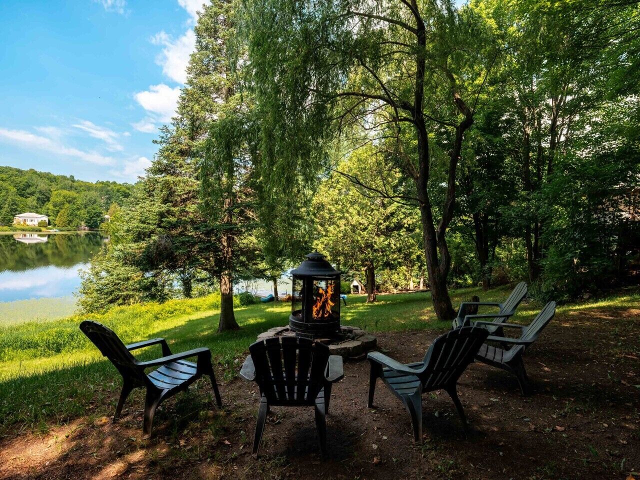 Un espacio al aire libre con sillas Adirondack y un hogar para fuego, rodeado de árboles y un lago.